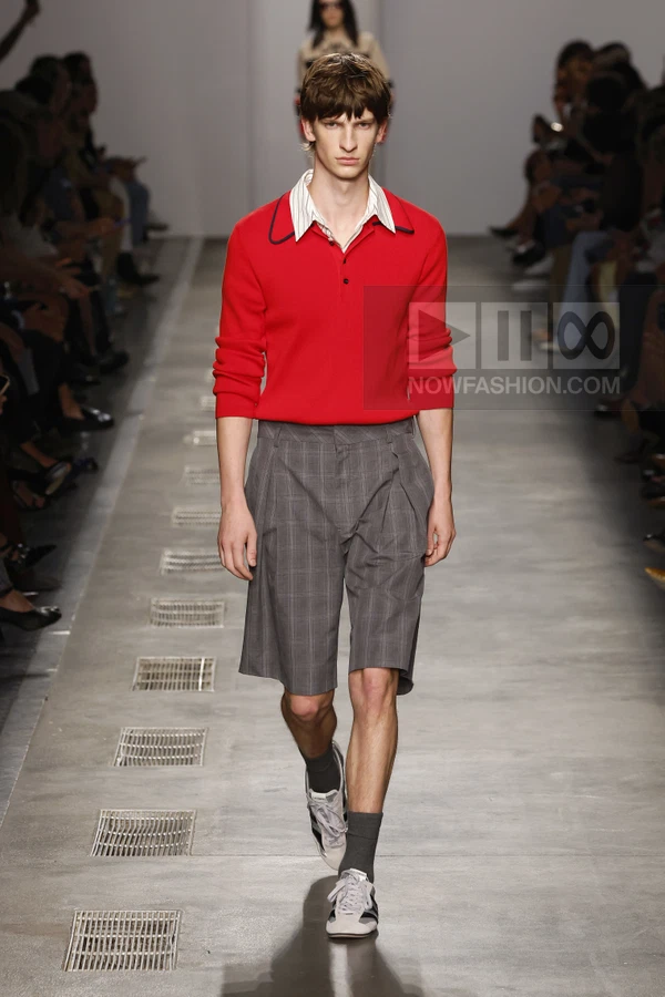 A person walking on a runway wearing a red polo shirt, gray plaid shorts, and sneakers with gray socks.