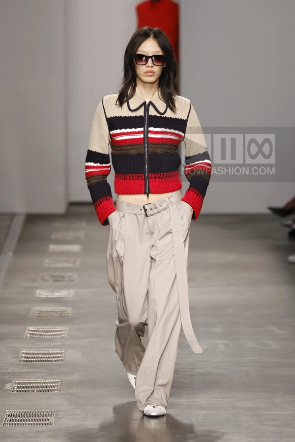 Model wearing a striped knit zip-up cardigan and beige wide-leg trousers on a runway.