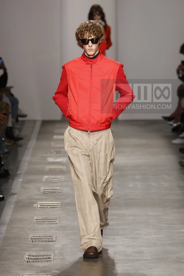 A person walks down a runway wearing a bright red zip-up jacket with ribbed sleeves and beige wide-leg trousers.