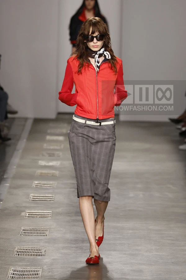 Model walking in a red zip-up jacket with ribbed sleeves, a gray plaid pencil skirt, white belt, and red flat shoes.