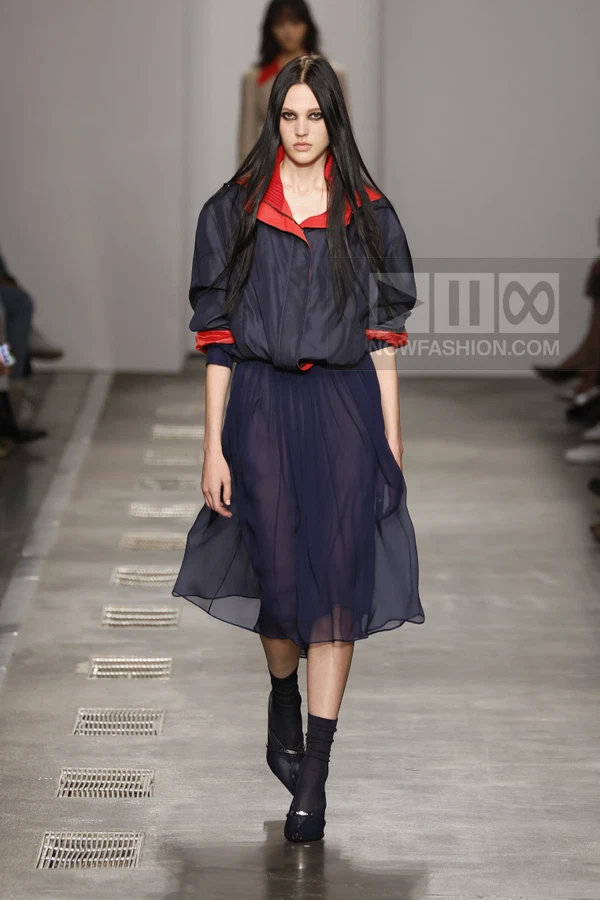 Model walking on runway wearing a navy blue windbreaker jacket over a sheer navy midi skirt with red collar details.