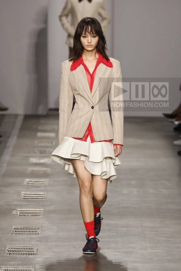Model walking in a beige plaid blazer, red shirt, and white tiered mini skirt with navy sneakers and red socks.