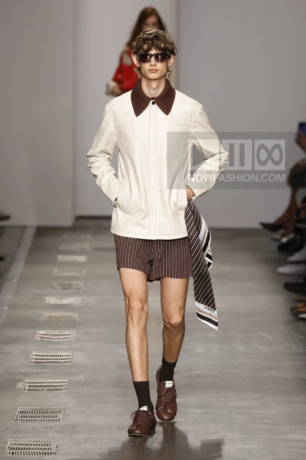 A person wearing a white leather jacket with a brown knit collar, striped shorts, and brown sneakers on a runway.