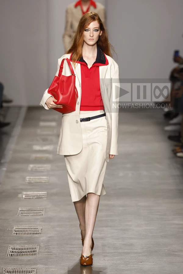 Model walking in a white blazer and skirt, red polo shirt, and red bucket bag on a runway.
