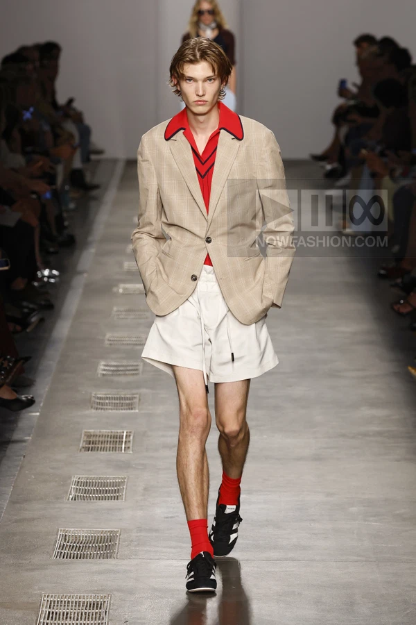 Model wearing a beige plaid blazer, red polo shirt, and white shorts with drawstring waist on a runway.