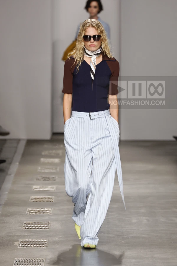 Model walking in a navy short-sleeve top, pinstriped wide-leg trousers, and a neck scarf on a runway.