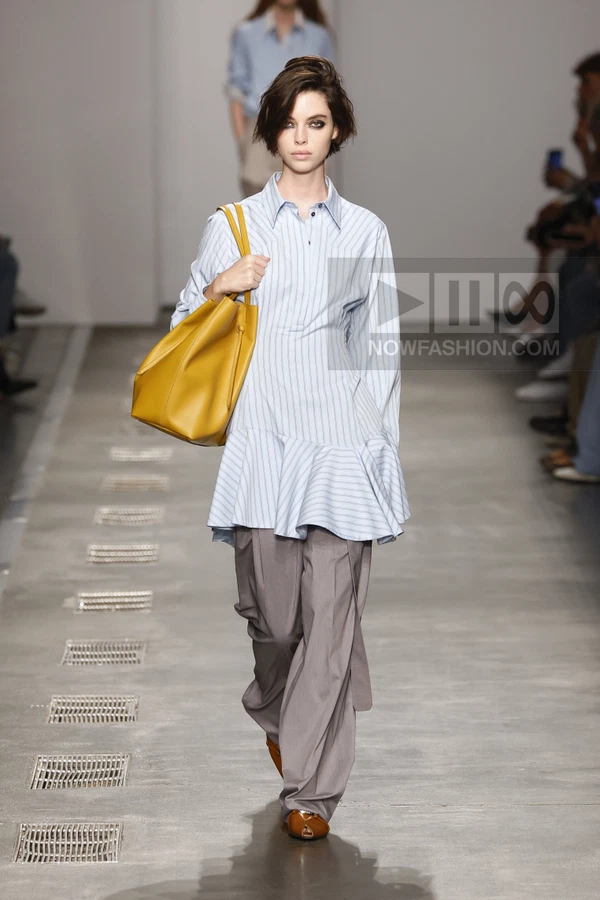 Model walking in a light blue striped shirt dress with a ruffled hem, gray wide-leg trousers, and a yellow shoulder bag.