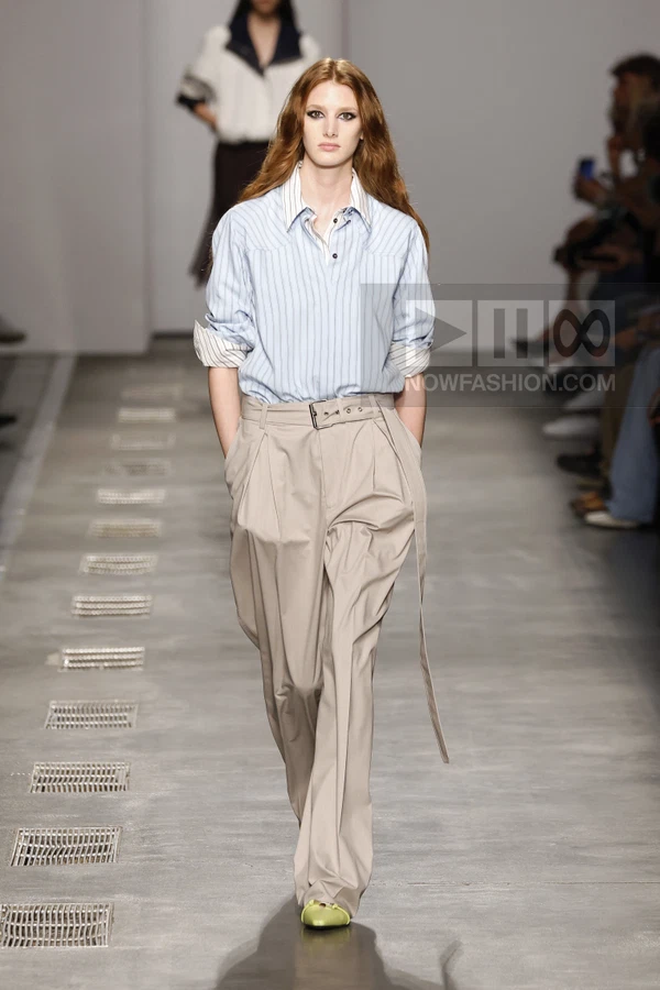 Model walking on a runway wearing a light blue striped shirt tucked into beige pleated trousers with a buckled belt.