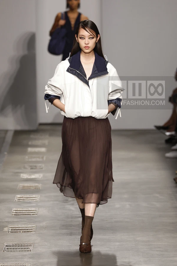 Model walking on runway wearing a white windbreaker jacket over a dark brown sheer midi skirt and brown socks with heels.