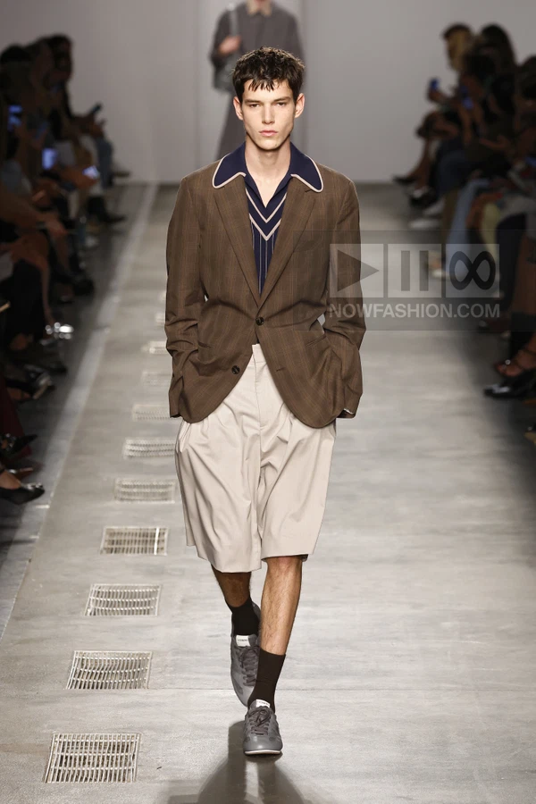 Model walking on runway wearing brown plaid blazer, navy v-neck shirt, and beige pleated shorts with gray sneakers.