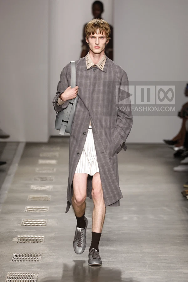 Model walking in a gray plaid trench coat over white pinstriped shorts, carrying a gray shoulder bag on the runway.