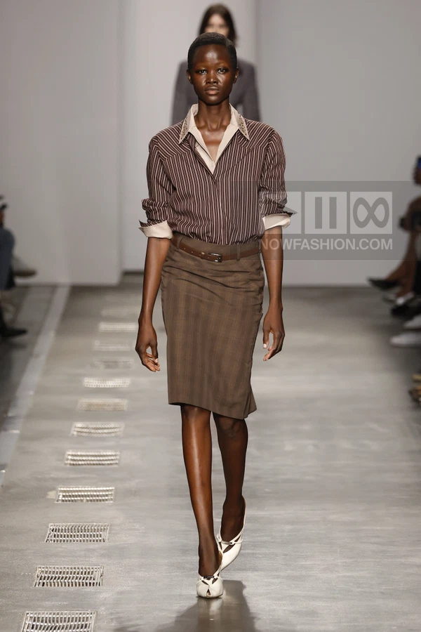 Model walking in a brown striped shirt, brown pencil skirt, and white pointed-toe shoes on a runway.