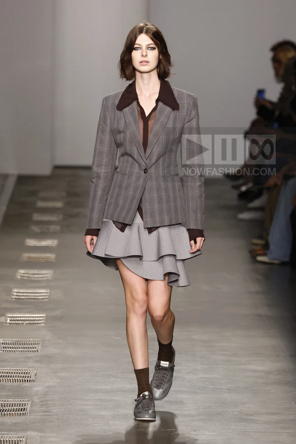 Model walking in a gray plaid blazer, sheer brown shirt, and tiered gray mini skirt with gray sneakers and brown socks.