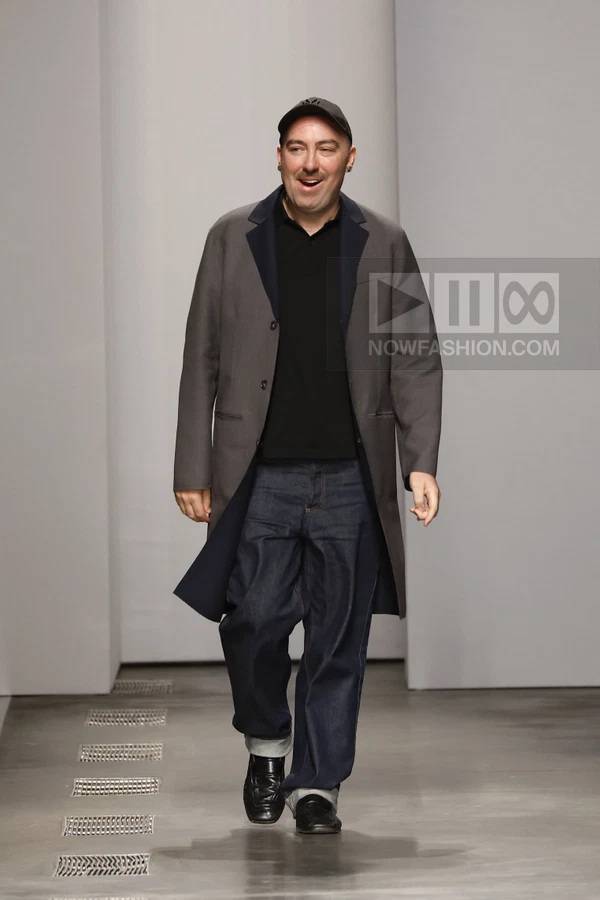 A person walking on a runway wearing a gray coat, black polo shirt, dark denim jeans, and a baseball cap.
