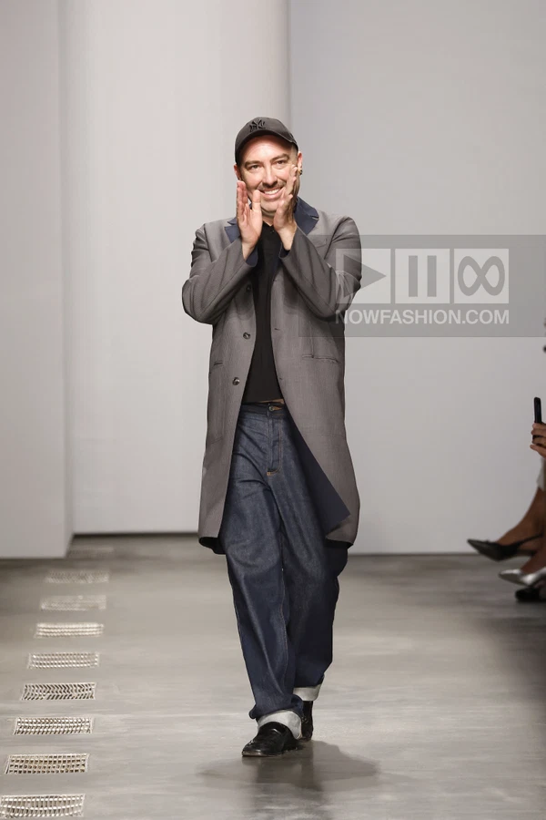 A person wearing a gray coat, black shirt, dark denim jeans, and a baseball cap walks on a runway, clapping hands.