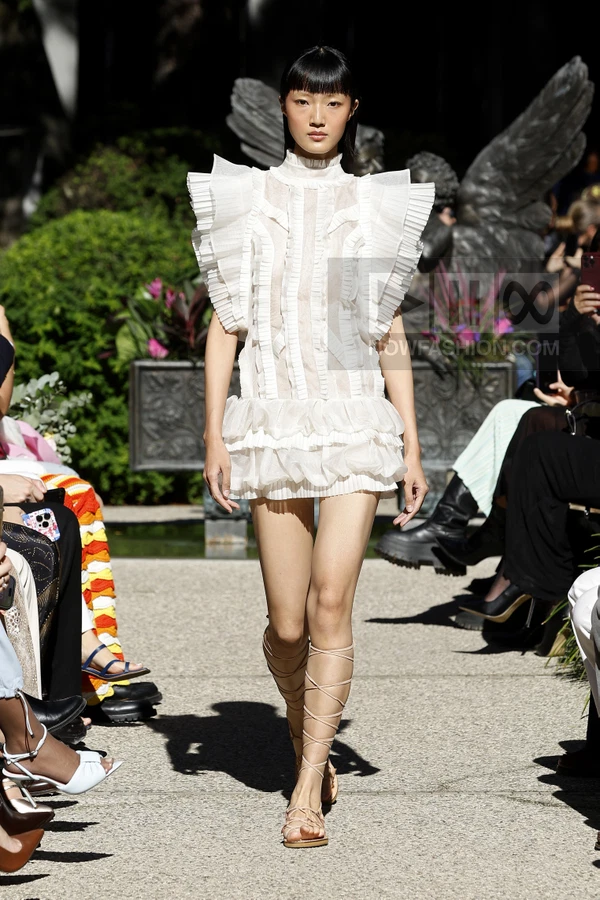 Model walking in a white mini dress with ruffled shoulders and pleated panels, paired with lace-up gladiator sandals.