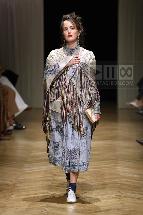 Person walking on a runway wearing a floral print dress, a fringed shawl, and white lace-up shoes with dark socks.