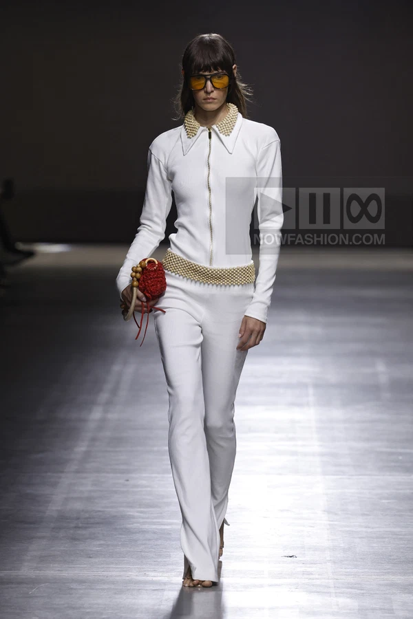 Model wearing a white zip-up jumpsuit with pearl collar and belt, carrying a small red crochet bag on a runway.