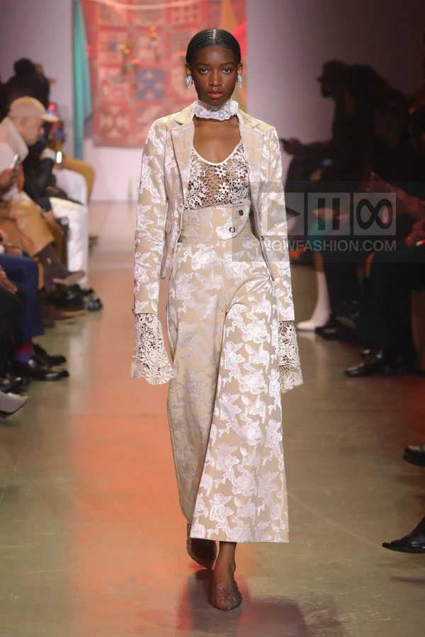 A model walks the runway wearing a beige and white brocade blazer and matching wide-leg trousers with lace details.