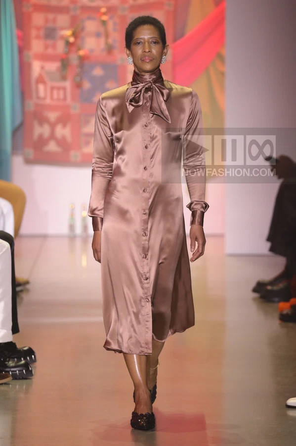 A person walks the runway in a long-sleeved, button-down satin midi dress in a muted mauve color with a neck tie.