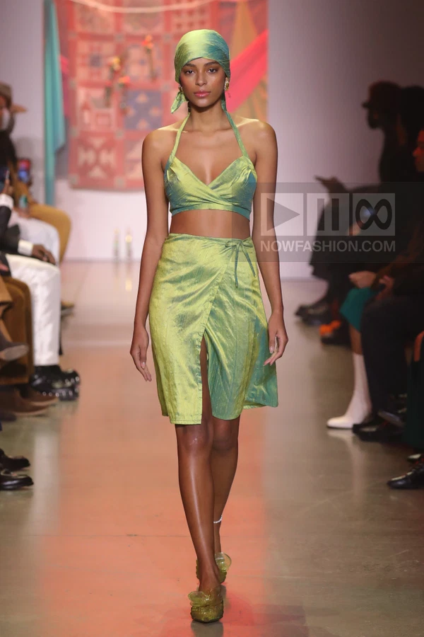 Model wearing a lime green two-piece set with a wrap skirt and headscarf on a runway.