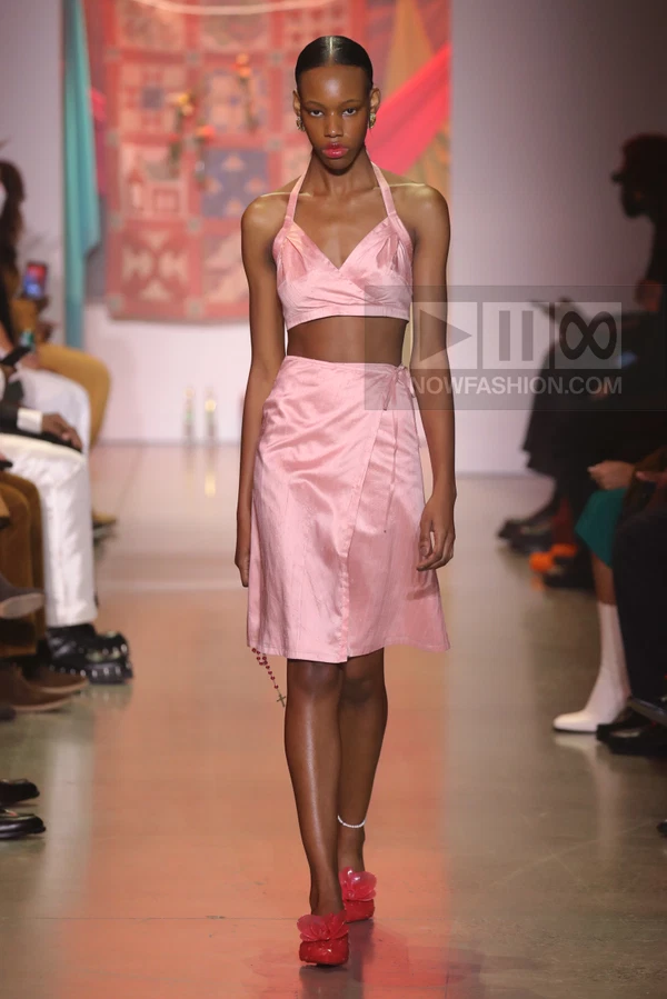 Model walking in a light pink satin two-piece set consisting of a halter crop top and a wrap skirt on a runway.