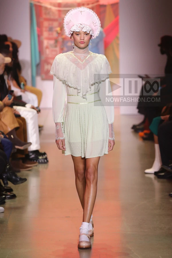 A model walks the runway wearing a pale green mini dress with a ruffled collar, lace cuffs, and a pink bonnet headpiece.
