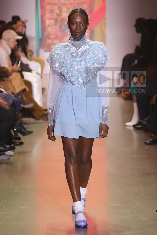 A model walks the runway wearing a light blue mini dress with a sheer floral embroidered capelet and matching mary janes.