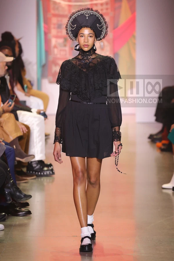 Model walks runway in black mini dress with lace yoke, ruffled collar, and ornate headpiece, paired with mary jane shoes.