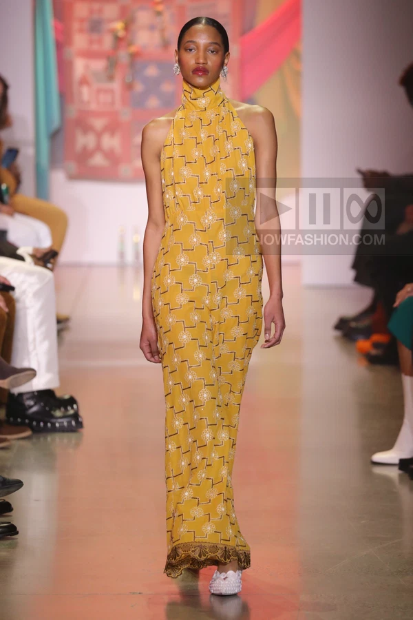 Model wearing a yellow halter-neck jumpsuit with white geometric floral embroidery and a dark brown scalloped hem.