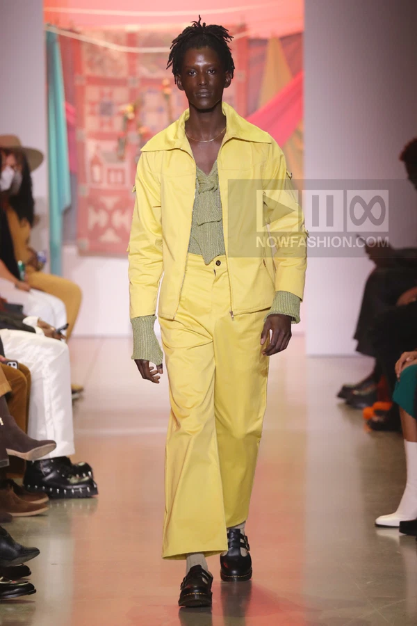 Model wearing a yellow jacket and matching wide-leg trousers with a textured green knit top on a runway.