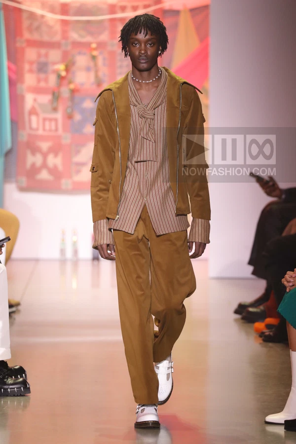 A person walks the runway wearing a brown jacket, striped shirt with a knotted collar, and matching brown trousers.