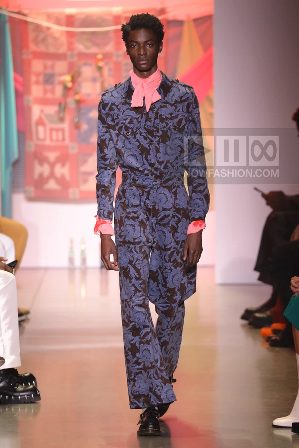 A person wearing a blue and brown floral print jumpsuit with a pink neck scarf and cuffs walking on a runway.