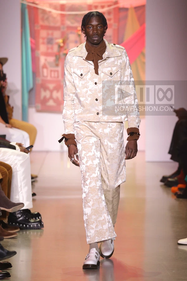 A person walks the runway wearing a cream-colored floral jacquard jacket and matching trousers with brown accents.