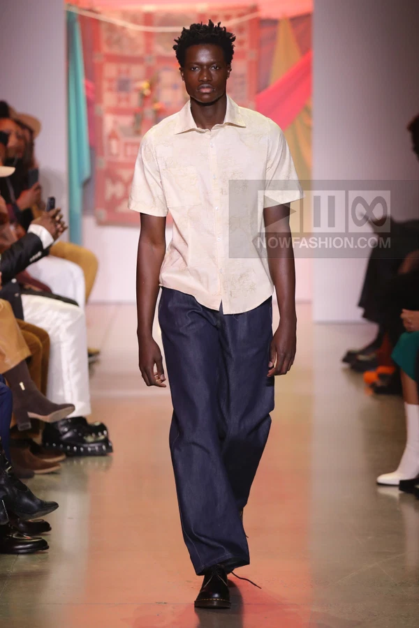 A person walks down a runway wearing a light-colored short-sleeve button-down shirt and dark blue wide-leg trousers.