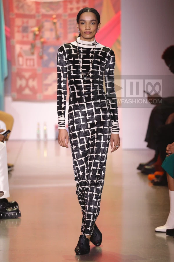 Model walking in a black and white grid-patterned long-sleeve jumpsuit with a high neck and black shoes.