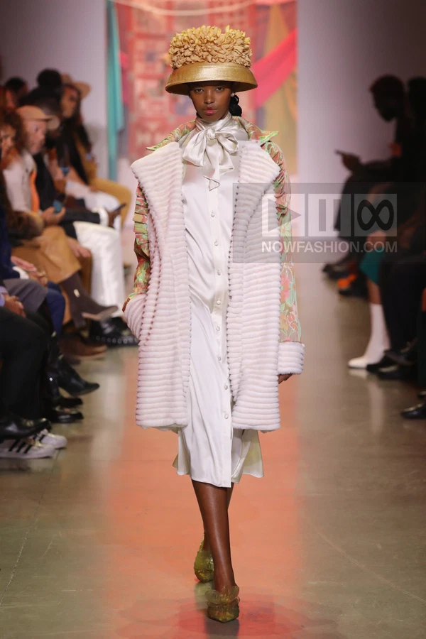 Model wearing a white satin shirt dress, a textured cream coat, and a floral hat walks down a runway.