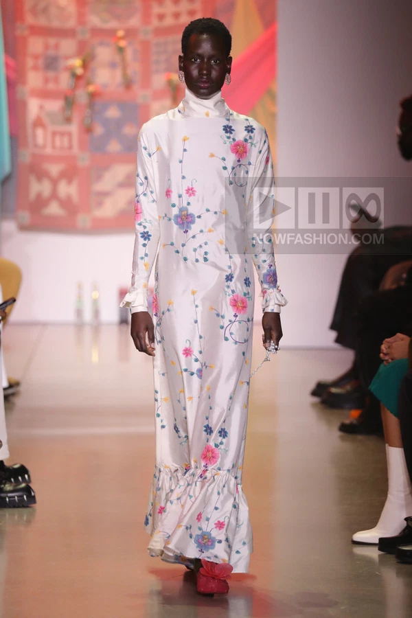 Model wearing a long-sleeved white silk dress with floral embroidery and ruffled cuffs and hem on a runway.