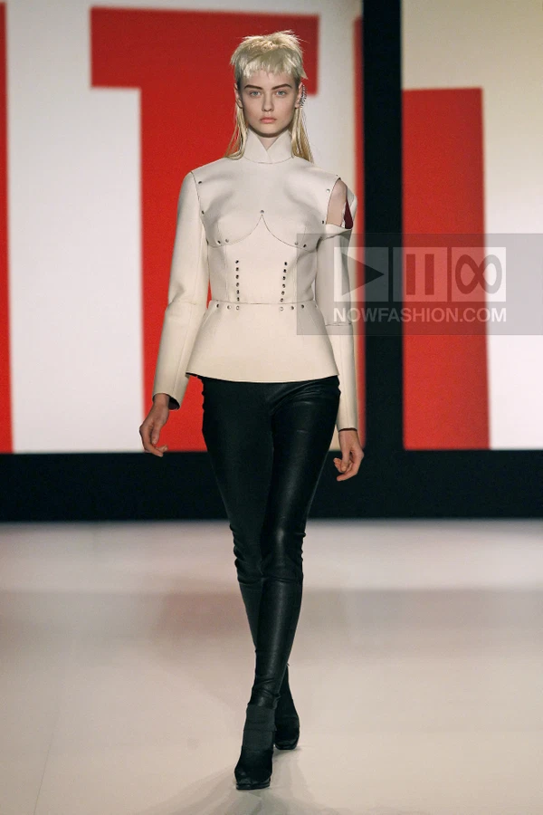 Model wearing a cream-colored structured top with metal studs and black leather trousers on a runway.
