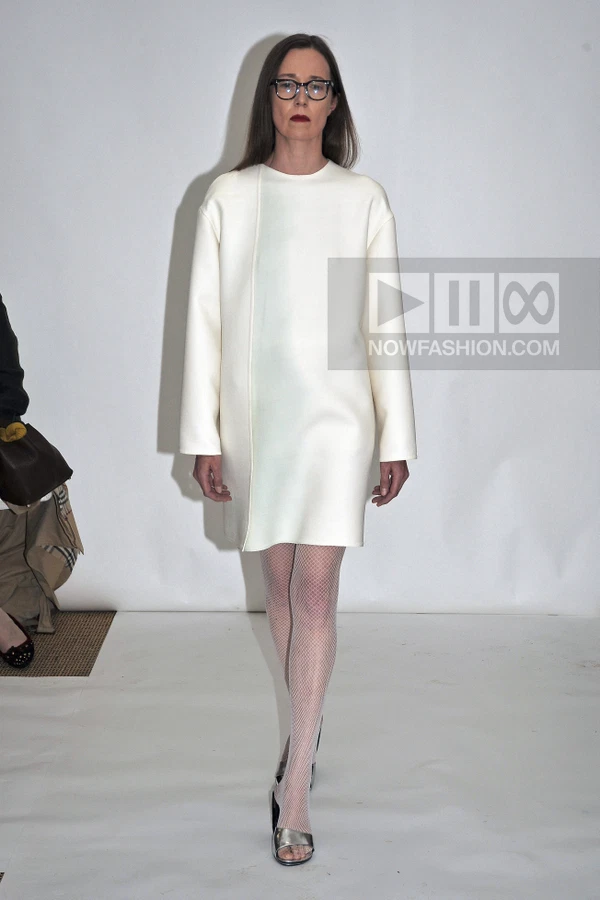 A person wearing a white shift dress and white fishnet tights walks on a white runway.