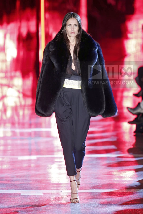 Model walking in a black fur coat, sheer black top, and dark tailored trousers on a red runway.
