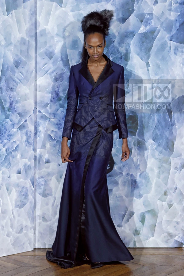 Model in a navy blue tailored jacket and floor-length skirt with lace-like patterns, standing against a blue backdrop.