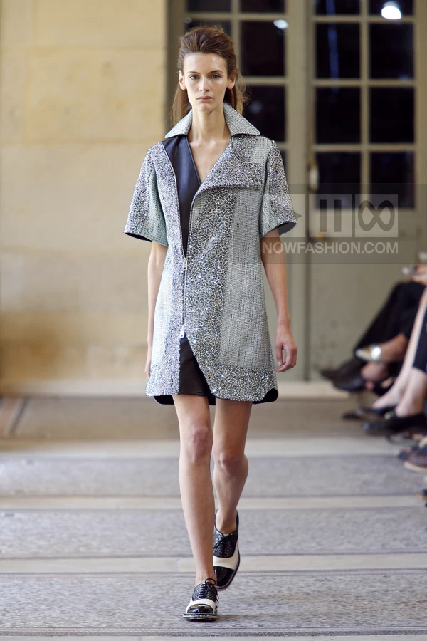Model walking in a short-sleeved, silver-textured coat dress with a front zipper, worn over black shorts and two-tone shoes.