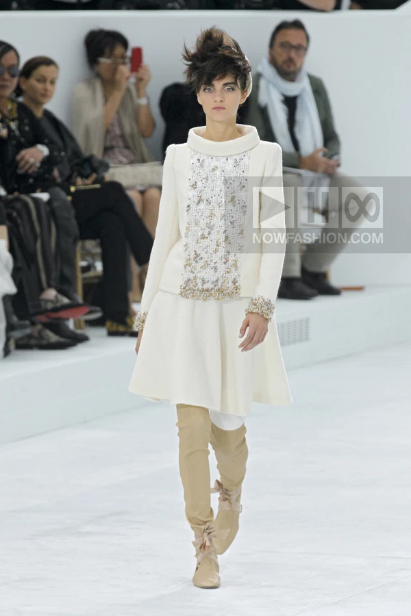Model walking in a white wool coat with embellished panel, white skirt, and beige over-the-knee boots on a runway.