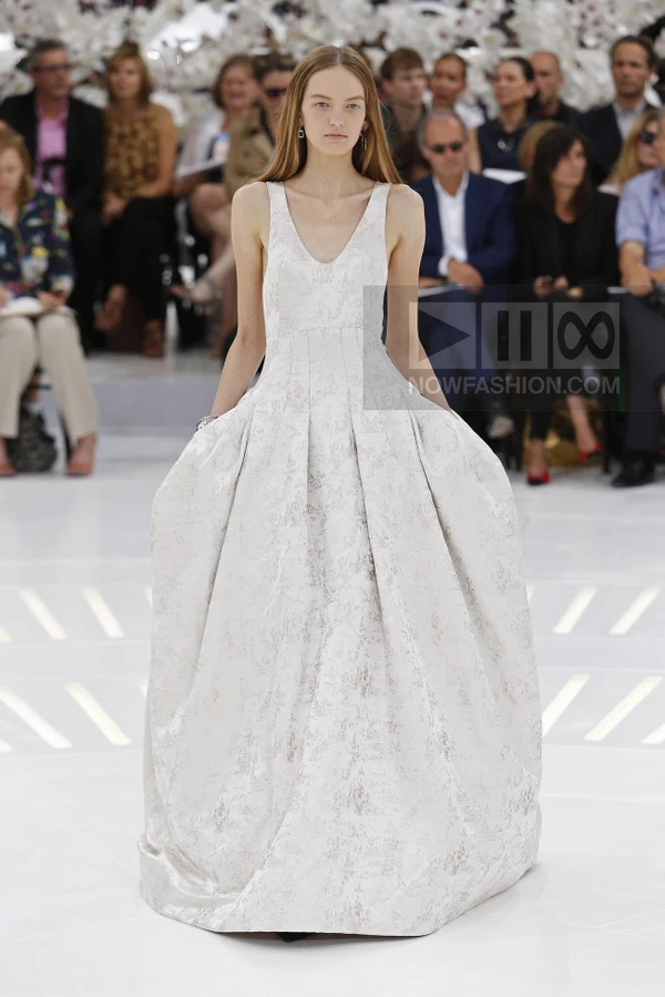 A model walks the runway wearing a white, floor-length, sleeveless gown with a full, pleated skirt and jacquard pattern.