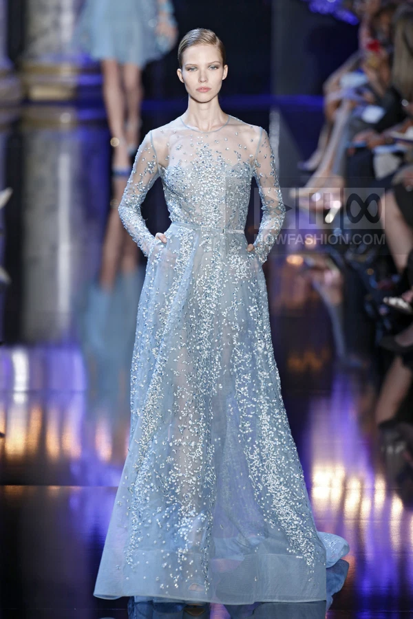 Model wearing a light blue floor-length gown with long sleeves and dense silver sequin embellishments on a runway.