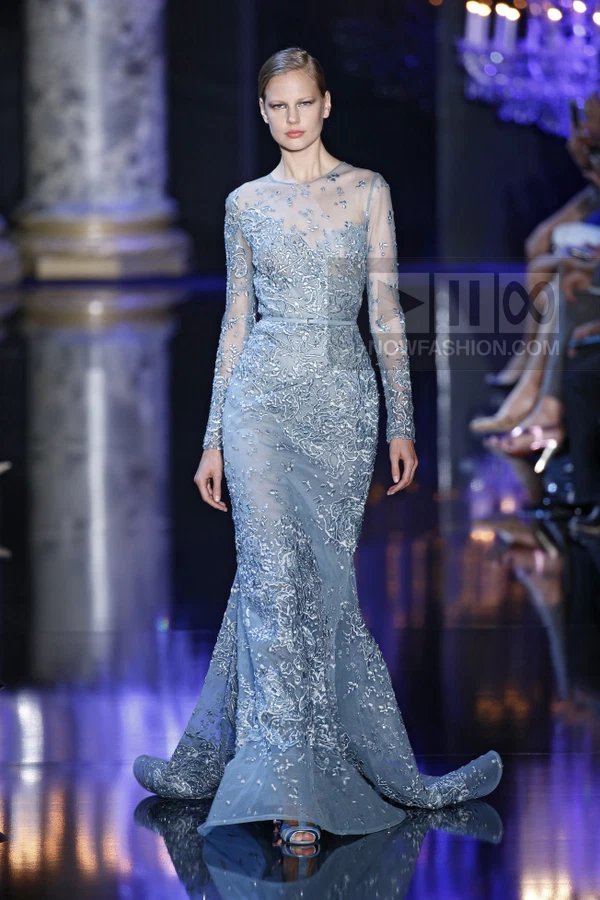 A model walks the runway in a floor-length, light blue, long-sleeved gown with intricate silver embroidery and a belt.