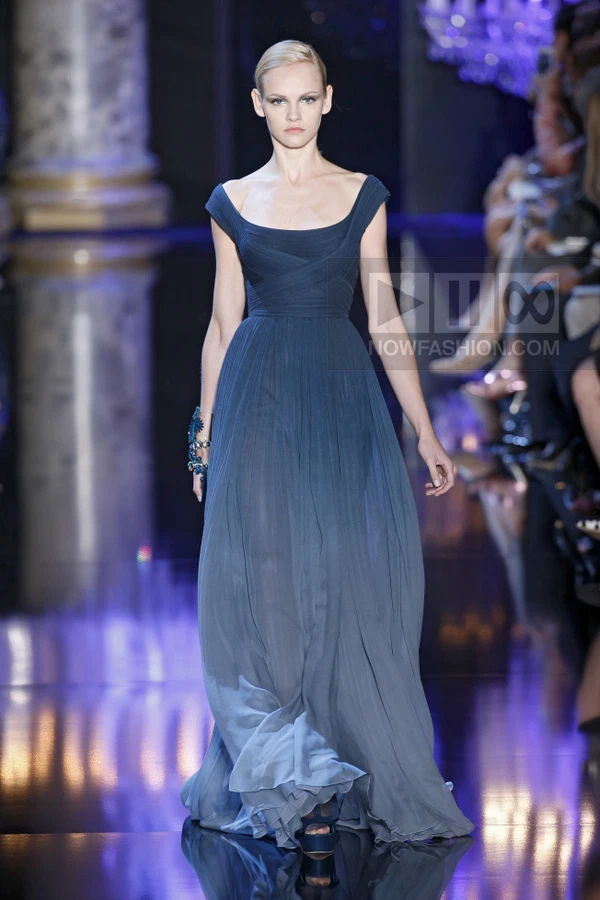 A model walks the runway in a floor-length, dark blue chiffon gown with a draped, fitted bodice and full skirt.