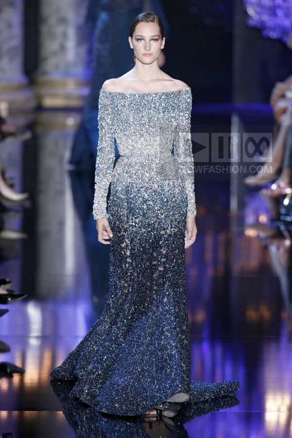 A model walks the runway in a floor-length, off-the-shoulder gown featuring an ombré sequin gradient from silver to navy.