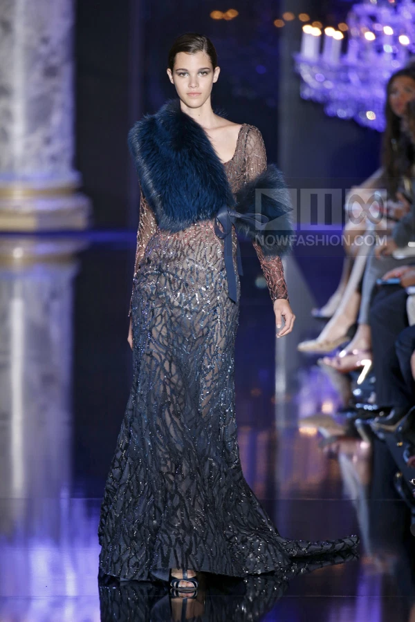 A model on a runway wearing a long, navy blue, sequined, sheer gown with a dark blue fur stole draped over one shoulder.